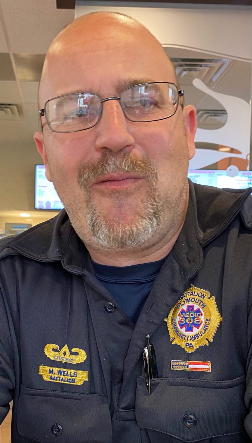 A middle-aged man with glasses and a trimmed beard smiles, wearing a dark uniform with badges that read "Battalion" and "Plymouth Medic EMS." He is indoors with a blurred background.