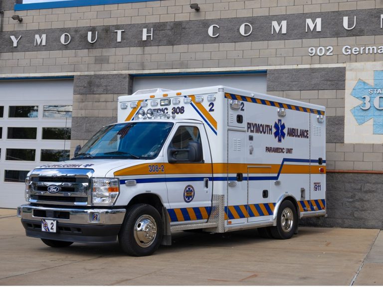 A white Plymouth Ambulance paramedic unit is parked outside a building with "Plymouth Community" on the wall. The ambulance displays blue and yellow stripes, emergency lights, and medical symbols.