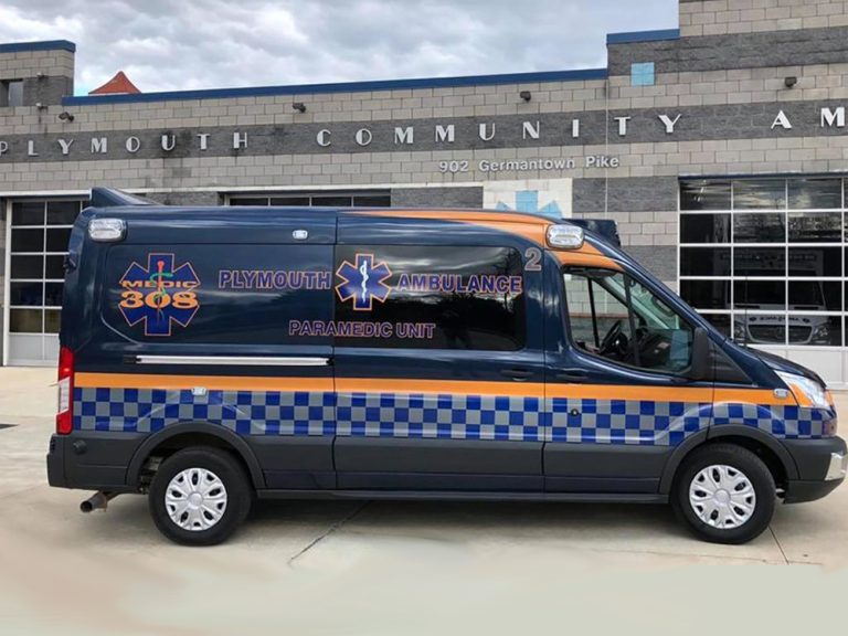 A blue and gray Plymouth Ambulance paramedic unit van is parked in front of a building with "Plymouth Community Amb" on the wall and "902 Germantown Pike" above the entrance.