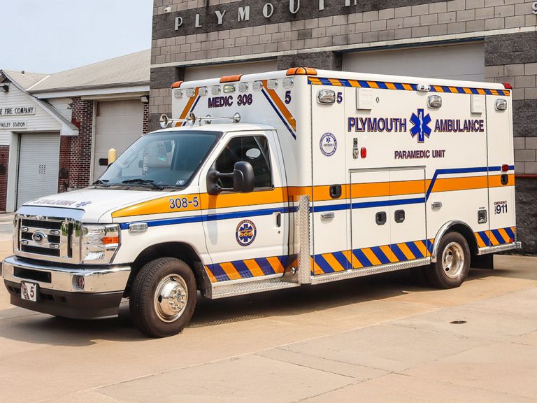A white Plymouth Ambulance paramedic unit is parked outside a building, featuring blue and orange stripes, emergency lights, and medical symbols.