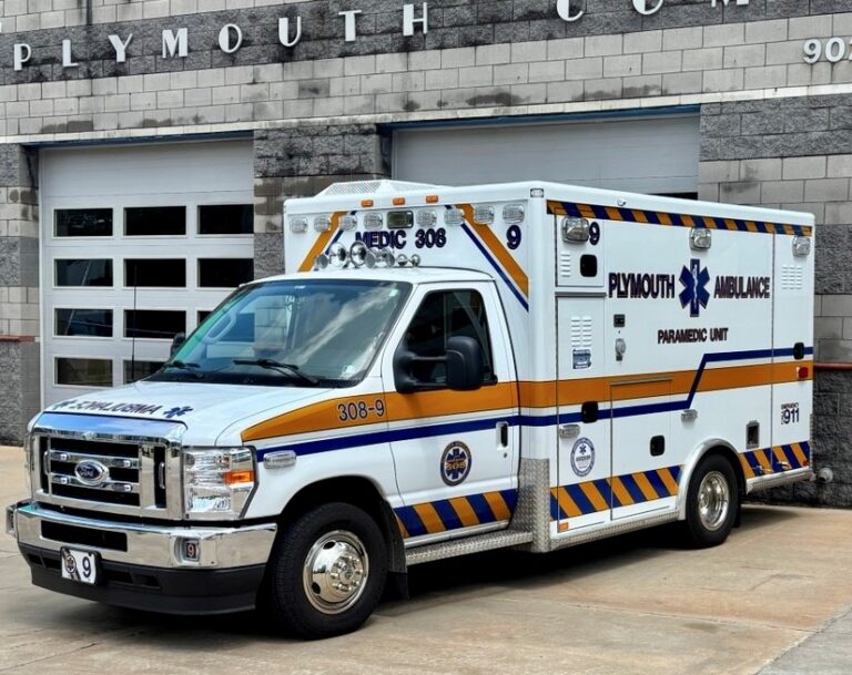 A white Plymouth Ambulance paramedic unit is parked outside a building with gray brick walls and garage doors. The vehicle has blue, orange, and gold stripes and emergency decals.