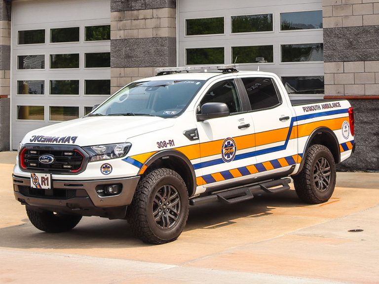 A white and yellow pickup truck marked "Paramedic" and "Ambulance" is parked outside a garage. The vehicle features emergency stripes, a medical emblem, and off-road tires.