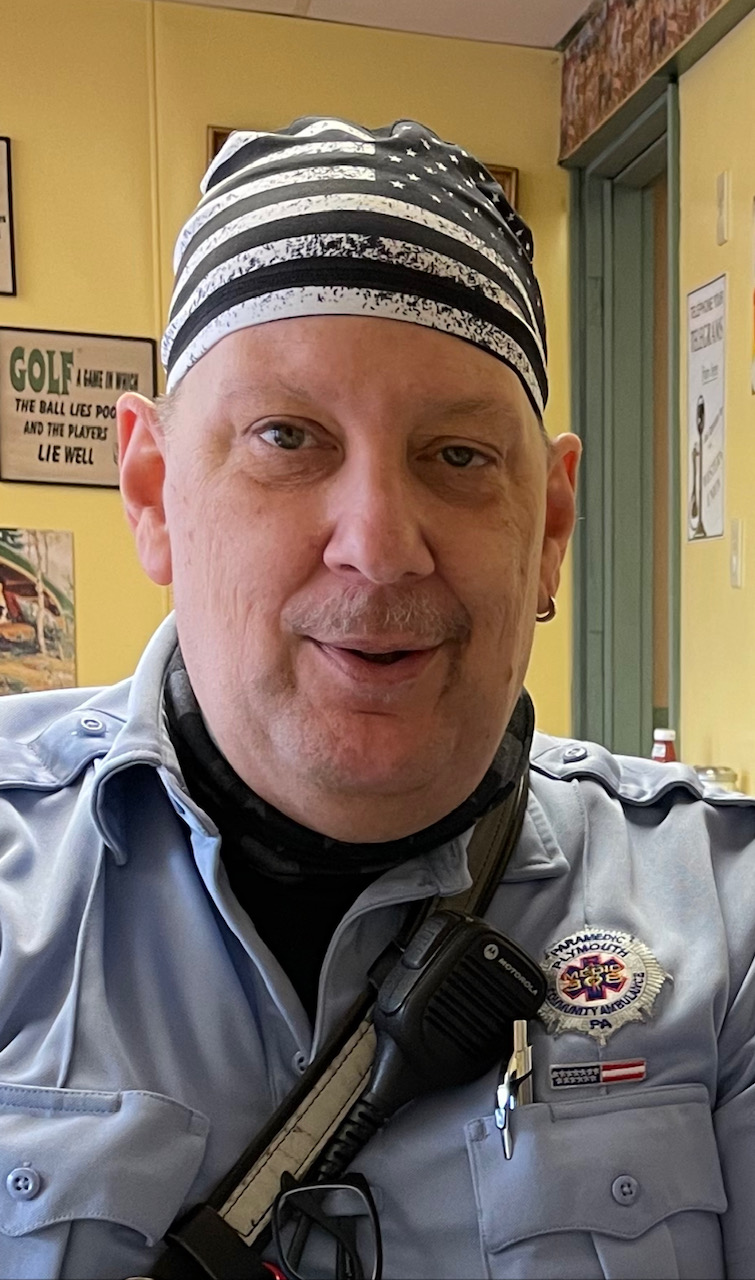 A person wearing a blue security uniform, a black-and-white striped bandana, and a radio sits indoors. The background includes posters and a doorway. The person is looking at the camera and smiling slightly.