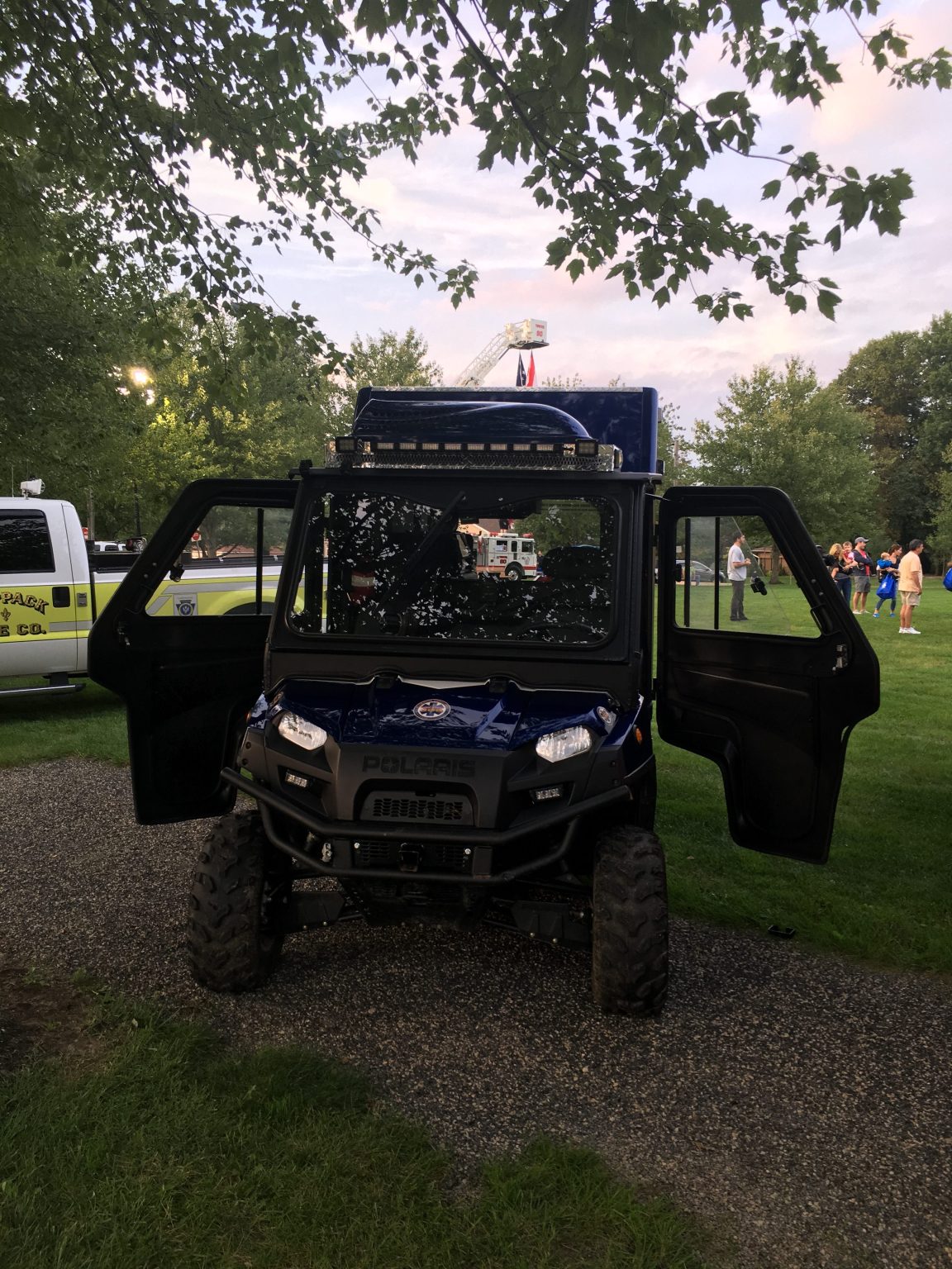 A blue police utility vehicle with both front doors open is parked on a gravel path in a park. People and emergency vehicles are visible in the background among green trees.