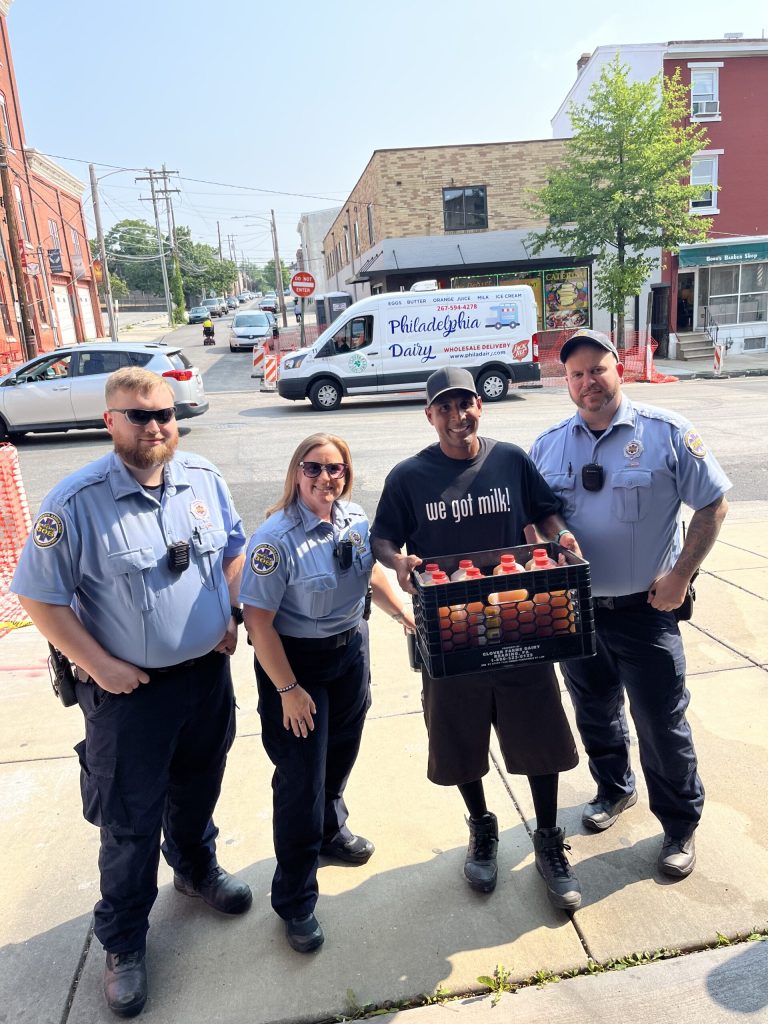 Four people stand on a sidewalk; three are in police uniforms, and one, wearing a "We got milk!" shirt and cap, holds a crate of milk cartons. A Philadelphia Dairy van is parked in the background.