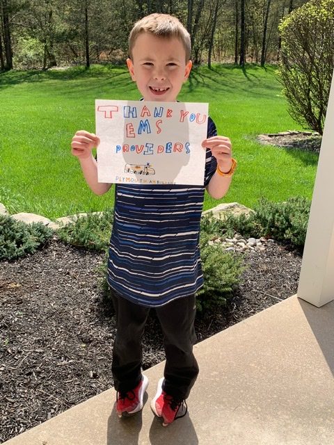 A smiling boy wearing a striped shirt and red sneakers stands outside on a sunny day, holding a colorful sign that says "Thank you EMS Providers" with a drawing of an ambulance.