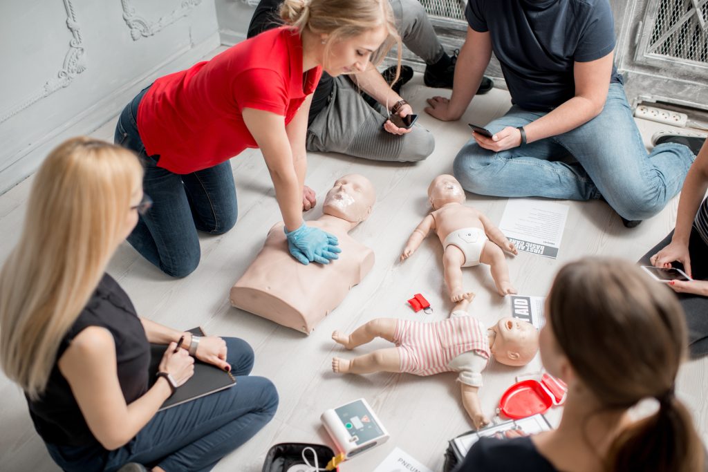 A woman in a red shirt demonstrates CPR on an adult mannequin while a group of people watches. Child and infant mannequins, medical equipment, and notepads are on the floor nearby.