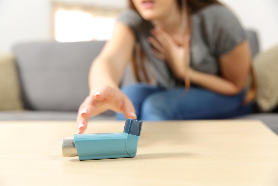 A woman sitting on a couch reaches urgently for an inhaler on a table, with one hand on her chest, suggesting she may be experiencing breathing difficulties.