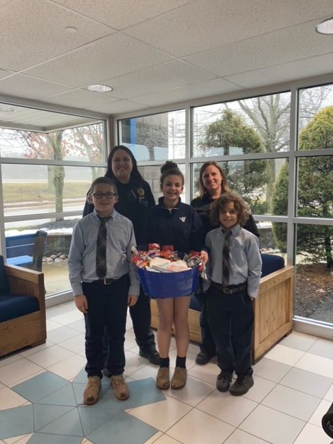 Three children in dress shirts and ties stand indoors holding a blue basket filled with snacks, while two smiling adults in uniforms stand behind them. Large windows show trees and overcast skies outside.