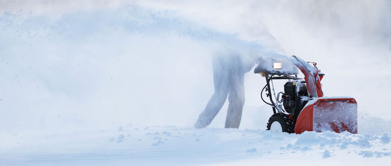 A person uses a red snow blower to clear a path through deep snow, with snow spraying high into the air and partially obscuring the person. The background is snowy and wintry.