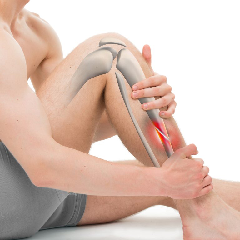A person holds their lower leg in pain, with an illustration overlay showing leg bones and a highlighted red area on the shin, indicating injury or inflammation.