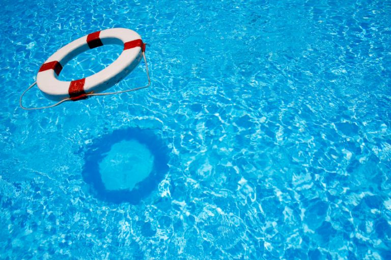 A white and red lifebuoy floats on the surface of a clear blue swimming pool, casting a distinct shadow on the water below.