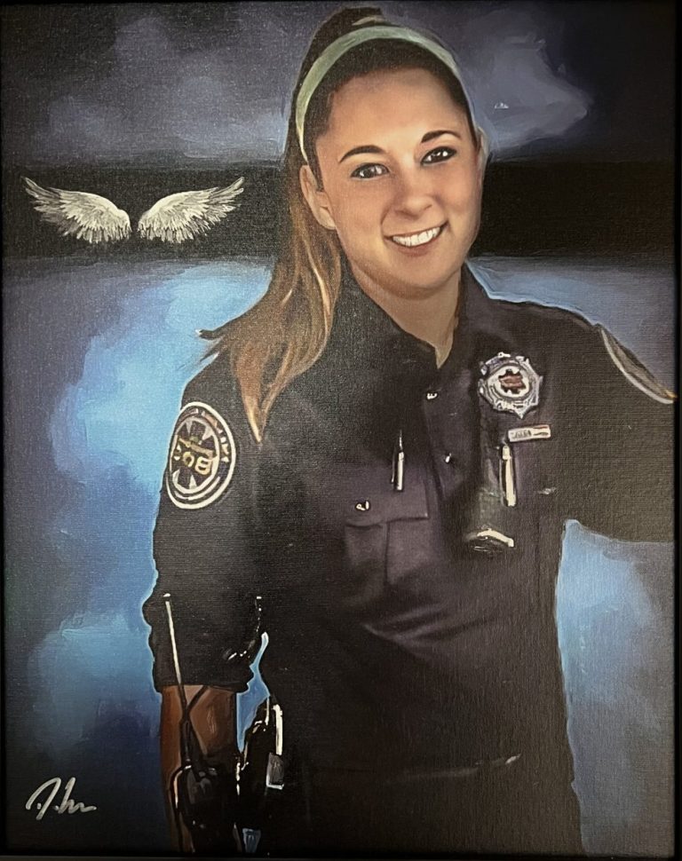 A smiling woman in a police uniform stands in front of a blue background with white angel wings behind her. She has long hair pulled back with a headband and wears a badge and police equipment on her belt.