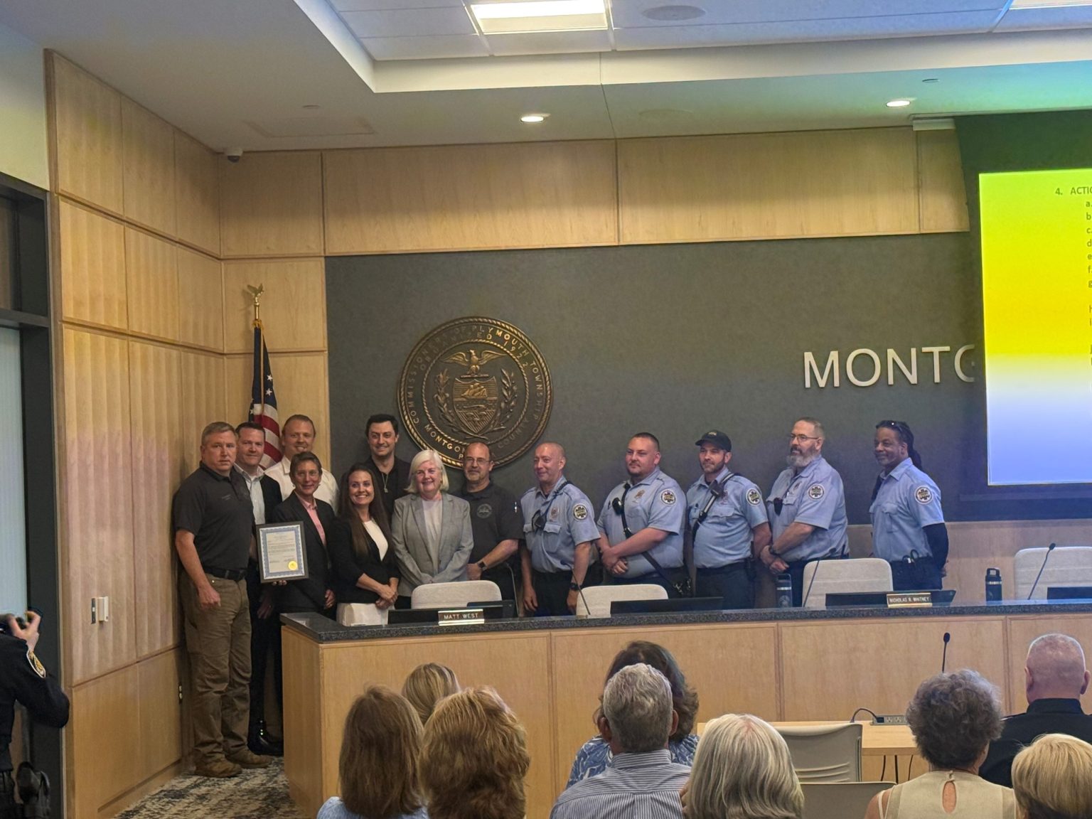 A group of people, including uniformed officers and civilians, pose together in a government meeting room in front of a seal and American flag. Some are holding a framed certificate while others smile at the camera. An audience is seated in front.