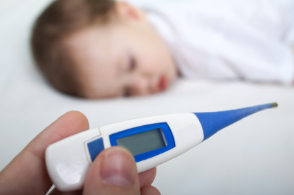 A close-up of a digital thermometer held in front of a blurred image of a baby lying down, suggesting the baby may be ill or have a fever.