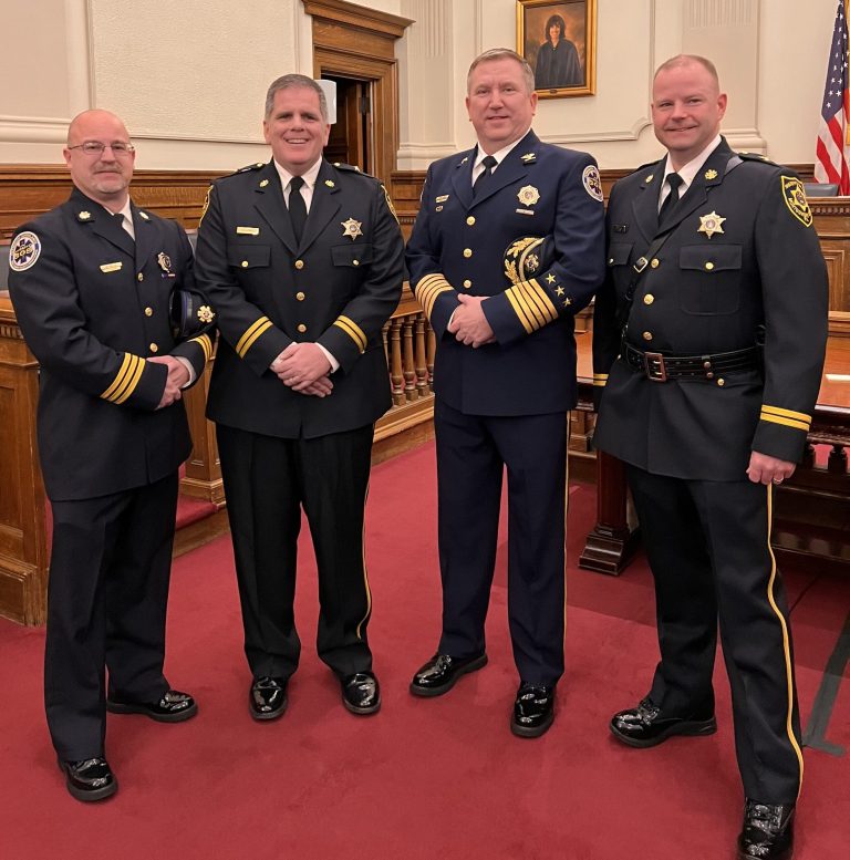 Four men in formal police uniforms stand smiling in a courtroom with wood paneling, a portrait on the wall, and an American flag in the background.