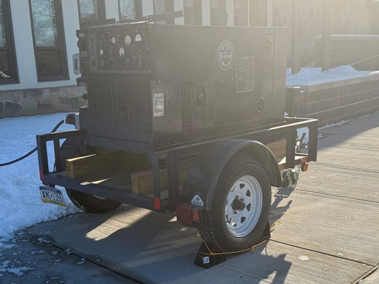 A large black generator mounted on a two-wheeled trailer is parked on a snowy concrete driveway, with control panels and dials visible on its side. Snow is piled along the edge of the driveway.