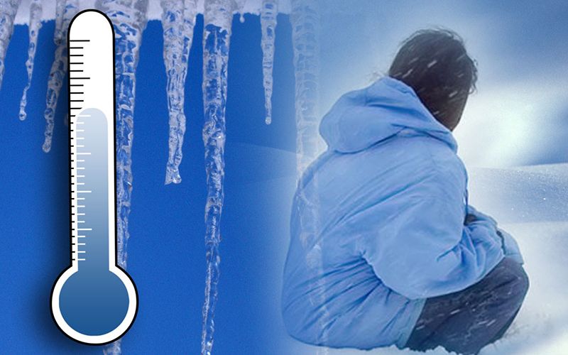 A person in a blue winter coat sits in snow under a cloudy sky, with icicles above and a thermometer showing very cold temperatures on the left.