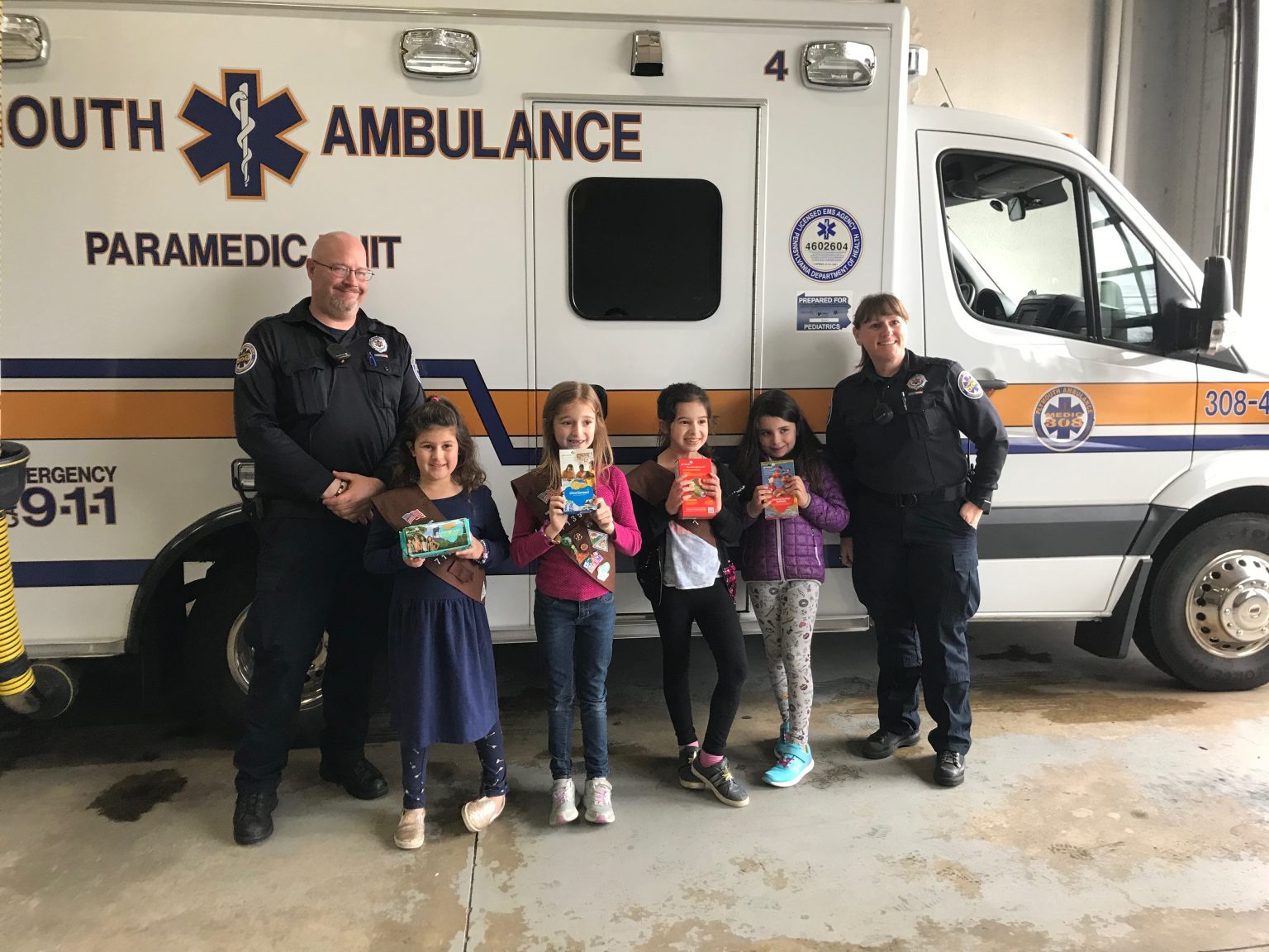Four children holding boxes of Girl Scout cookies stand in front of an ambulance with two paramedics, all smiling for the photo inside an emergency vehicle station.