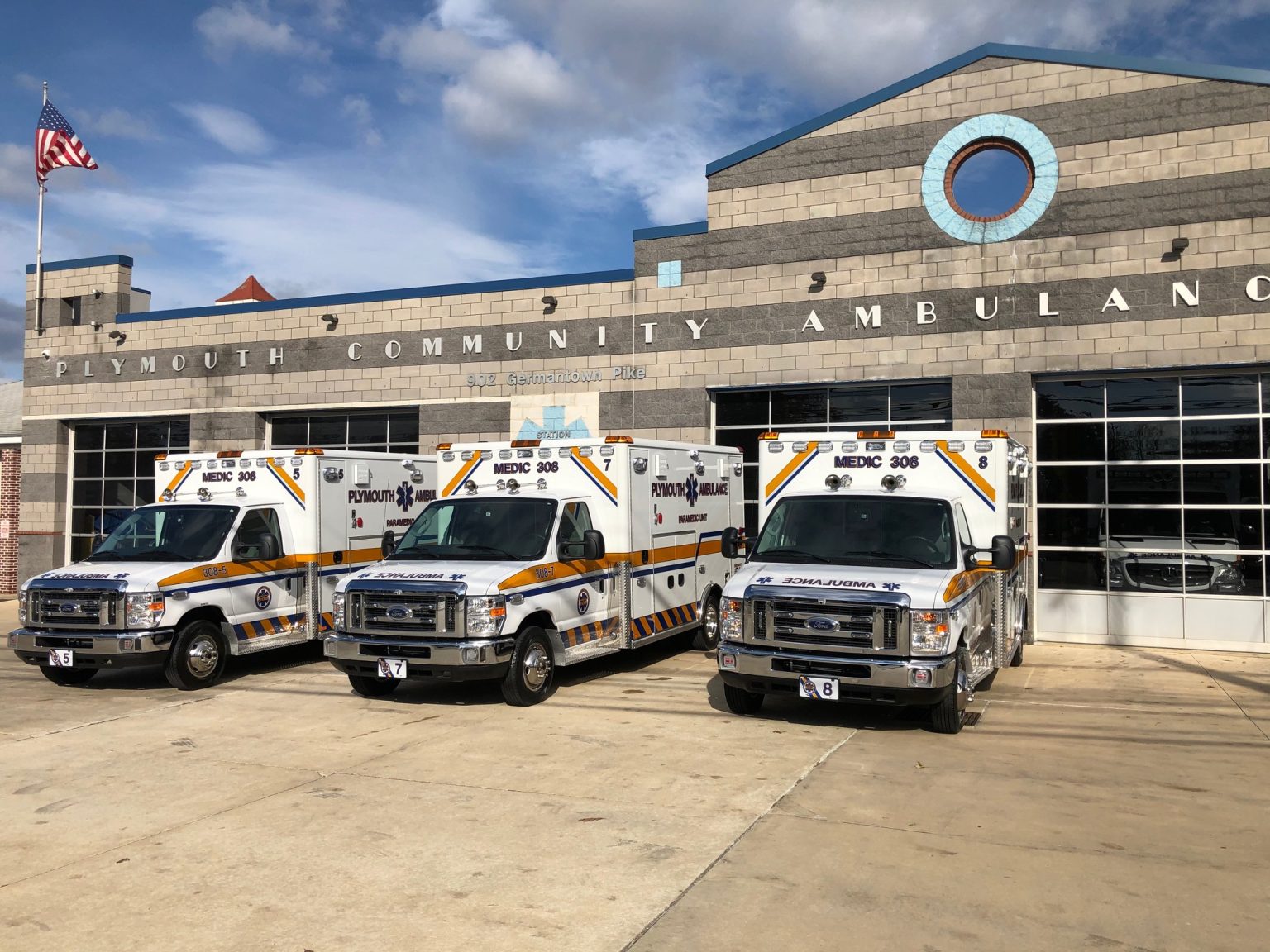Three ambulances are parked in front of the Plymouth Community Ambulance Association building on a sunny day, with the American flag flying to the left.