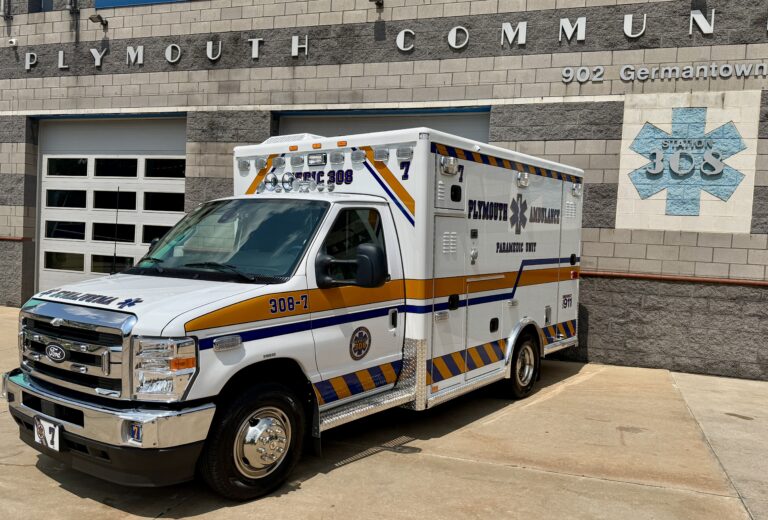 A white Plymouth ambulance with blue and yellow stripes is parked outside a building labeled “Plymouth Community” and “Station 308,” with an address number 902 Germantown visible on the wall.