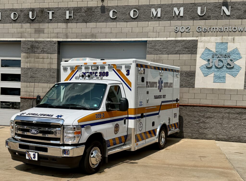 A Plymouth Ambulance vehicle is parked in front of a building with "Station 308" and "902 Germantown" visible on the wall. The ambulance has blue and yellow stripes and emergency markings.