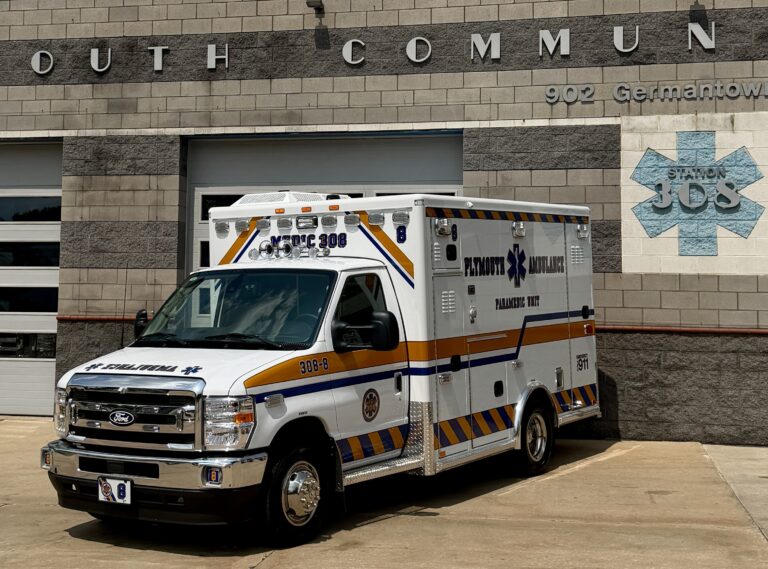 A white and yellow ambulance labeled "Medic 308" is parked outside a building with "Station 305" and a medical cross emblem displayed on the wall.