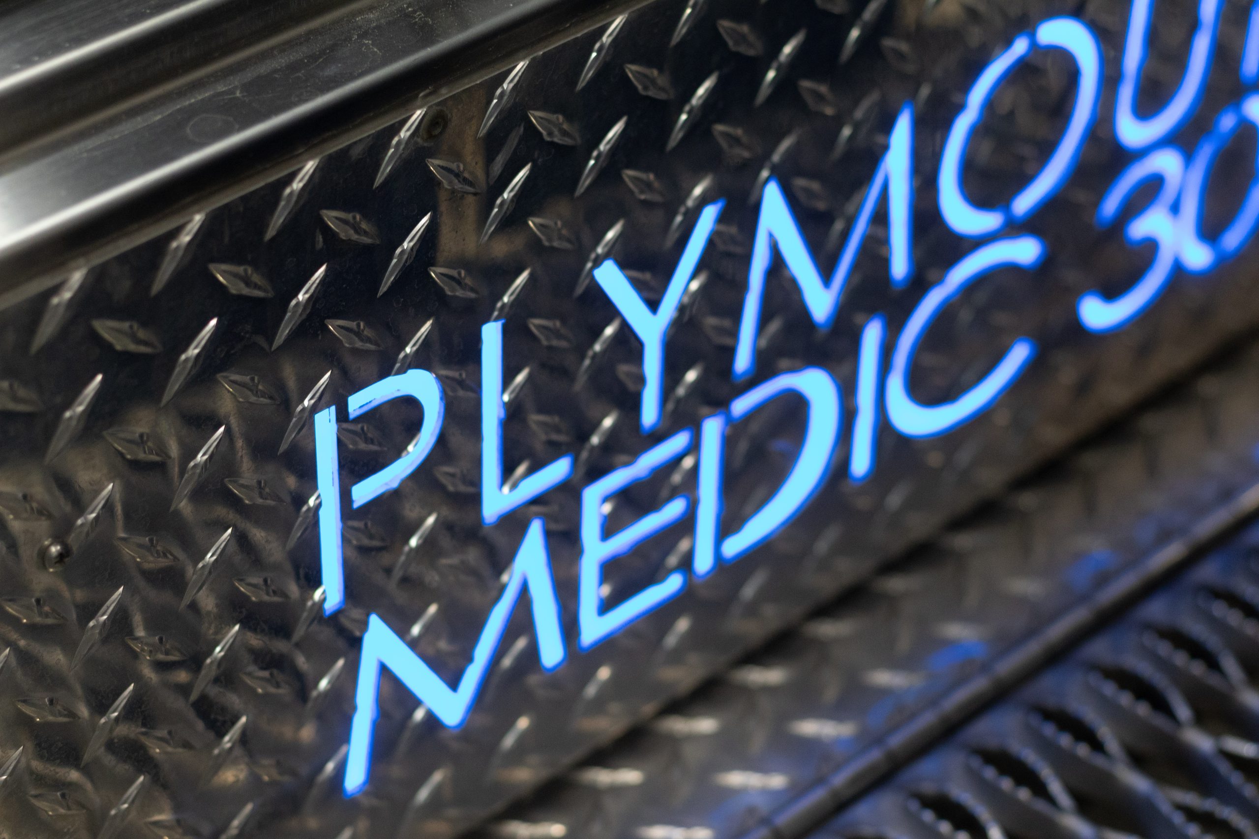 Close-up of metallic diamond plate with blue illuminated text reading "PLYMOUTH MEDIC 3" partially visible, suggesting emergency or medical vehicle signage.