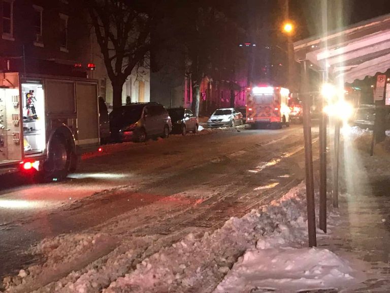 Fire trucks and emergency vehicles line a snowy, icy street at night with lights flashing. Cars are parked along the road, and snow piles up on the sidewalk. The scene suggests an emergency response in winter conditions.