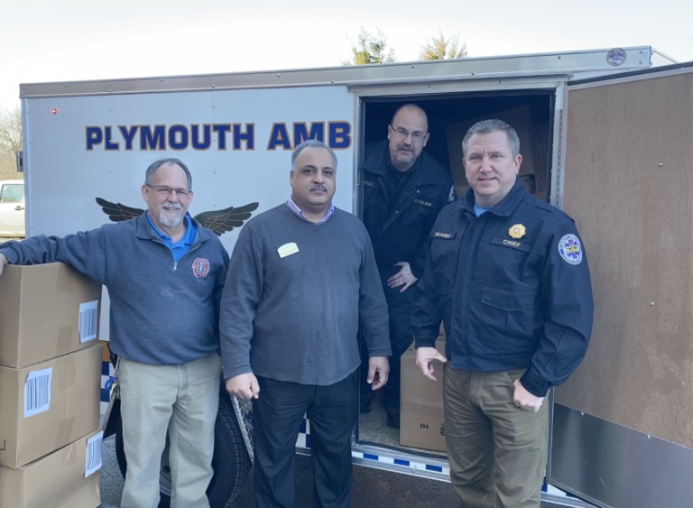 Four men stand smiling in front of a trailer labeled "PLYMOUTH AMB," with cardboard boxes stacked nearby, suggesting an organized supply or donation event.