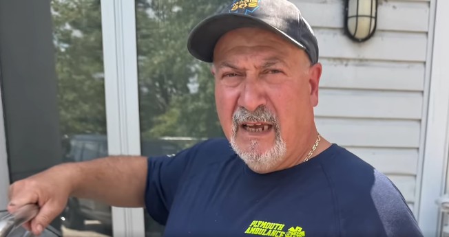 A middle-aged man wearing a navy cap and blue "Plymouth Ambulance" shirt stands outside by a light-colored wall, looking at the camera with a neutral expression.