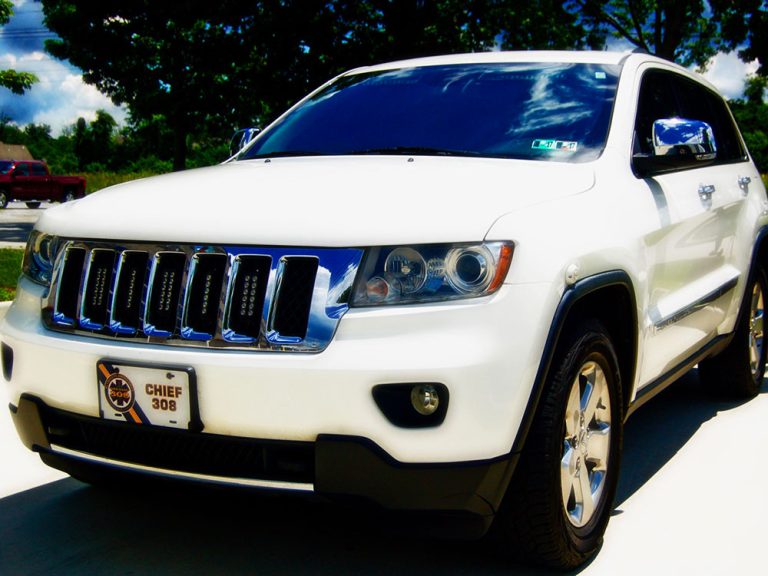 A white SUV parked on a driveway with trees and a red truck in the background. The front license plate reads "Chief 308." The sky is blue with some clouds.