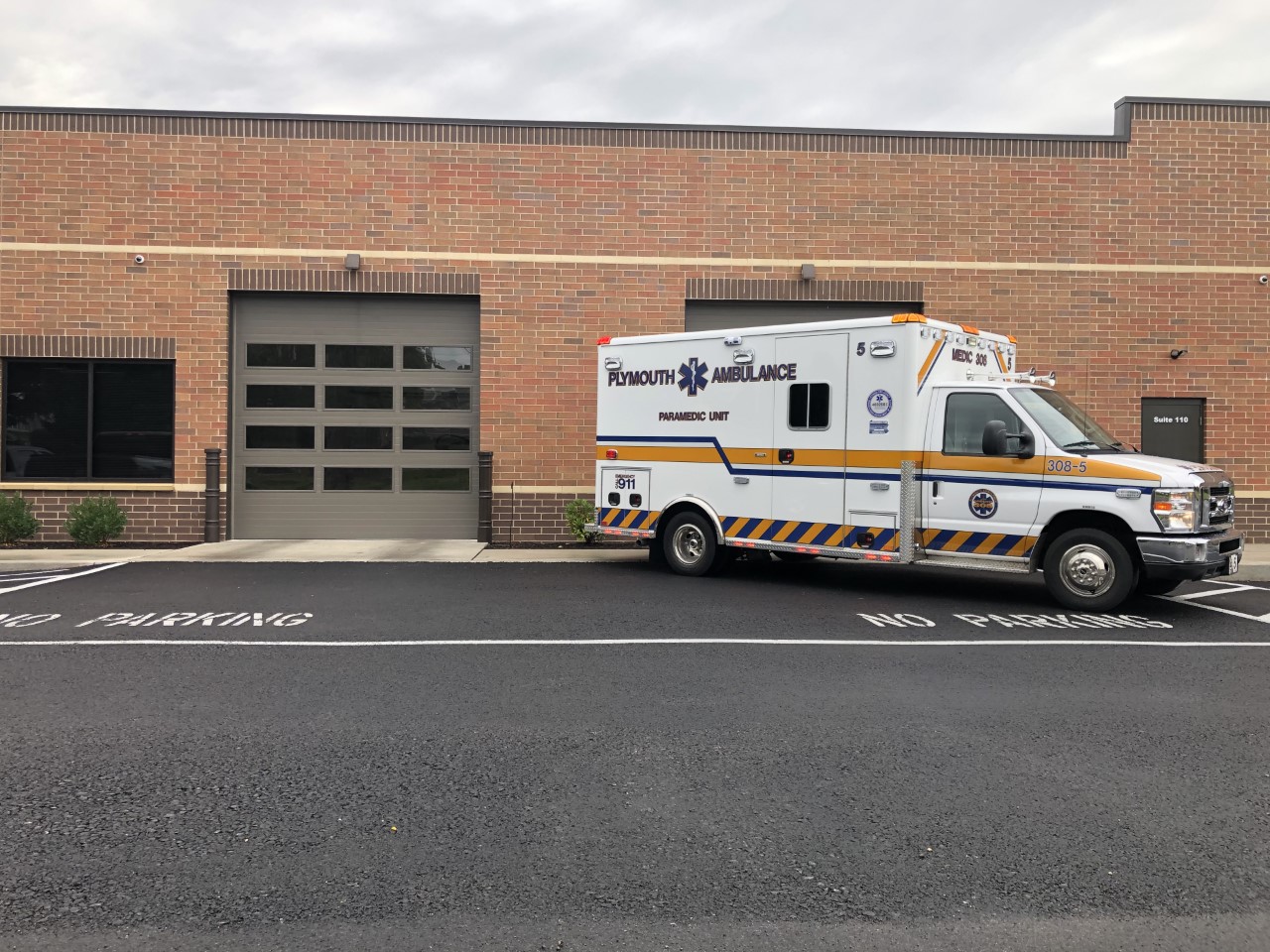 A white Plymouth Ambulance is parked in front of a brick building in a space marked "NO PARKING" on the ground near a garage door.