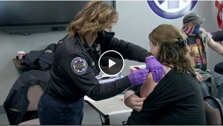 A healthcare worker wearing gloves and a mask administers a vaccine to a seated woman while another person waits nearby. The setting appears to be a clinic or vaccination site.