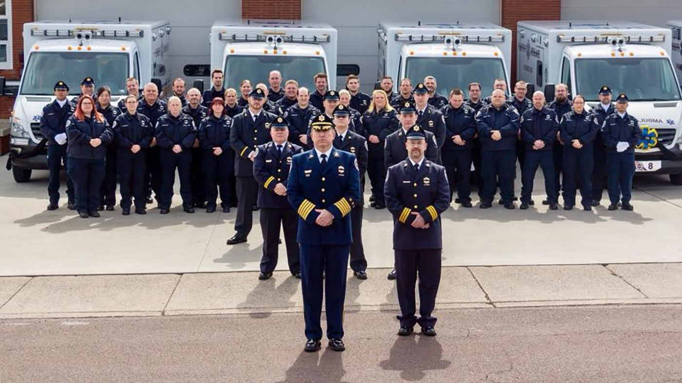 A large group of uniformed emergency medical personnel pose in front of several ambulances, with two officers standing prominently in front of the group.
