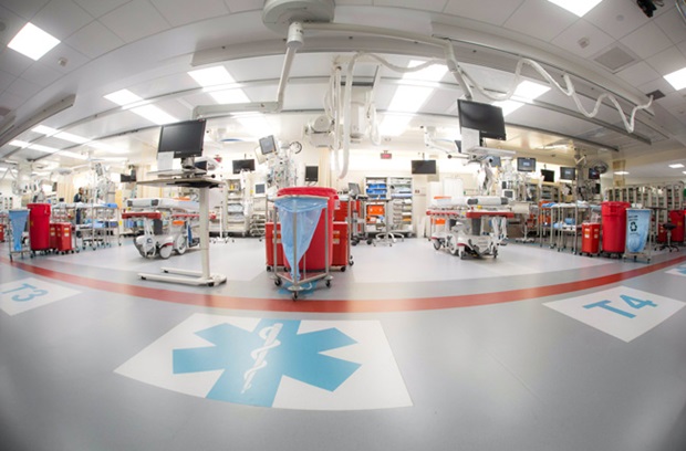 Wide-angle view of a modern hospital emergency room with multiple empty patient beds, medical equipment, monitors, and red supply carts. Large medical symbols and numbers mark sections on the clean, bright floor.