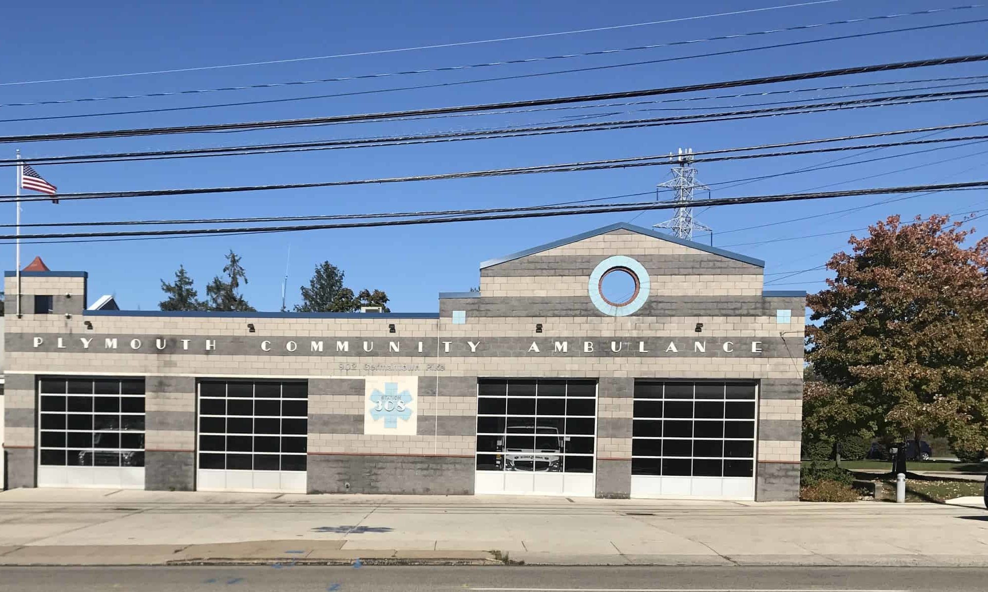 Gray brick building with three garage doors labeled "Plymouth Community Ambulance." An American flag is on a pole to the left, and a tree with autumn leaves is on the right. Power lines cross above the building.