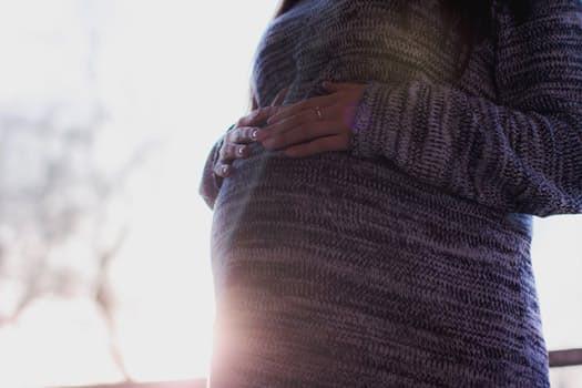 A person wearing a gray sweater stands with hands gently resting on their pregnant belly, with soft sunlight shining in the background.