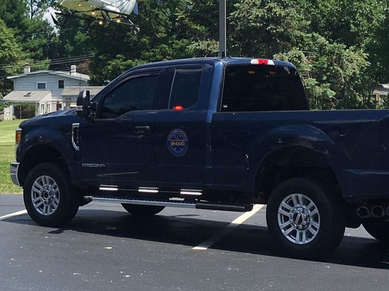 A dark blue pickup truck with tinted windows and a police or official emblem on the door is parked in a lot on a sunny day, with trees and houses in the background.