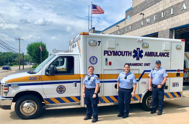 Three paramedics in uniform stand in front of a Plymouth Ambulance parked outside an ambulance station, with the American flag flying above the building in the background.
