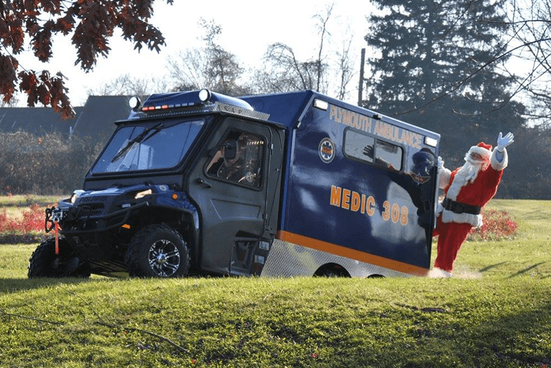 Santa Claus waves while stepping out of a blue Plymouth Ambulance vehicle labeled "MEDIC 908," parked on grassy terrain with trees and houses in the background.