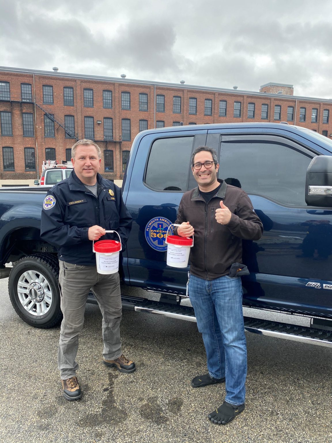 Two men stand next to a dark blue truck, each holding a red and white donation bucket. One man gives a thumbs up. Both are smiling. A brick building and cloudy sky are visible in the background.