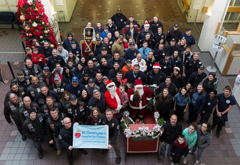 A large group of people, including firefighters, hospital staff, and people in festive costumes, gather in a hospital lobby around Santa Claus and Mrs. Claus, with a Christmas tree and a banner for St. Christopher's Hospital for Children.