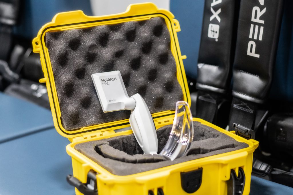 A McGRATH MAC video laryngoscope and two blades are securely placed in a yellow protective carrying case with custom foam inserts, positioned on a blue surface with black seat belts in the background.