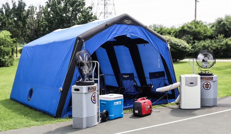 A large blue outdoor tent is set up on pavement, with portable fans, a generator, a cooler, and an air conditioning unit positioned outside. Lush green grass and trees are in the background.
